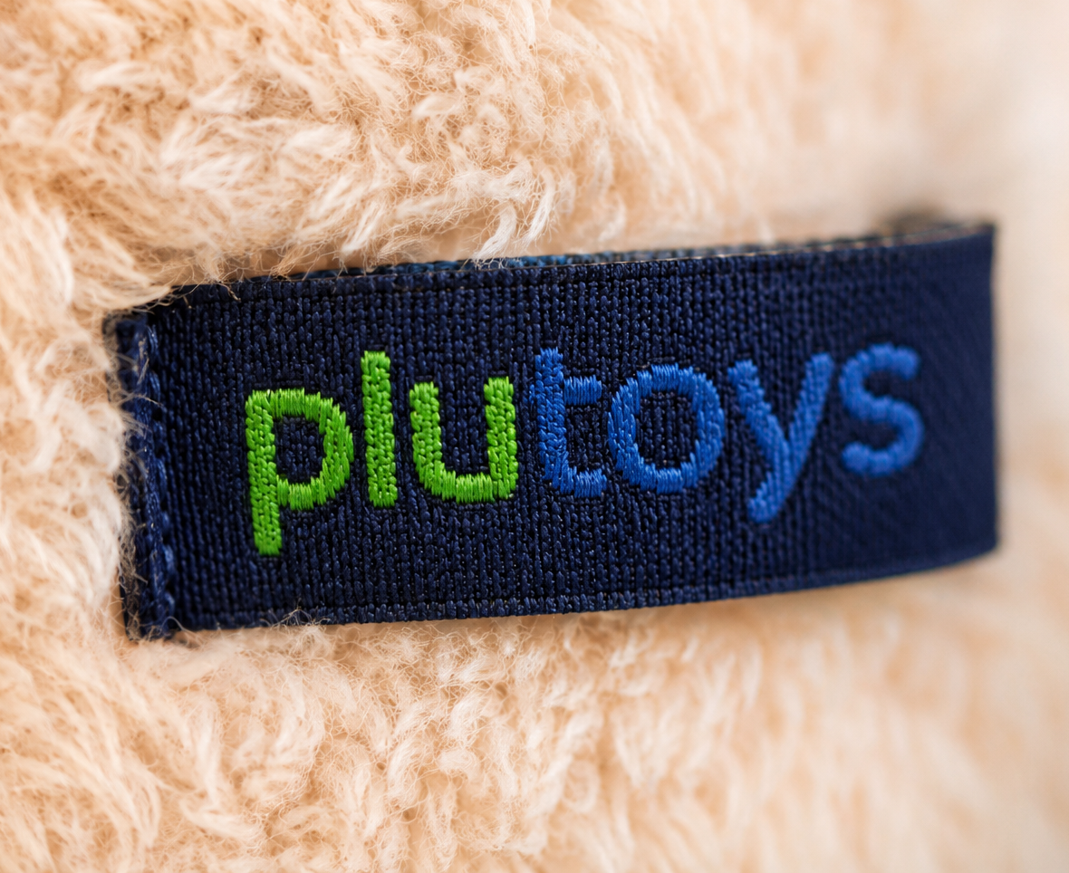 Ingenaaid Plutoys-label in donkerblauw garen op zachte crèmekleurige pluche